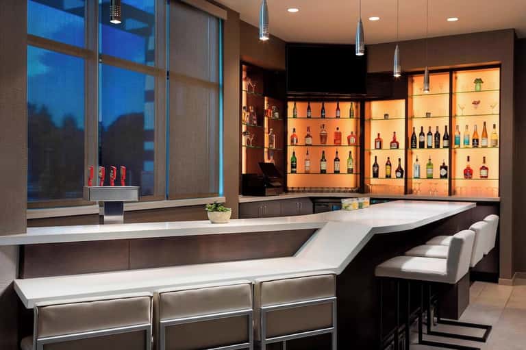 Upscale hotel bar with illuminated bottle display, white counter seating, and modern design