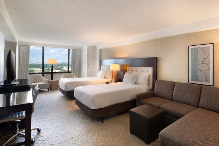 Modern hotel room with two beds, city view windows, and contemporary brown and beige decor