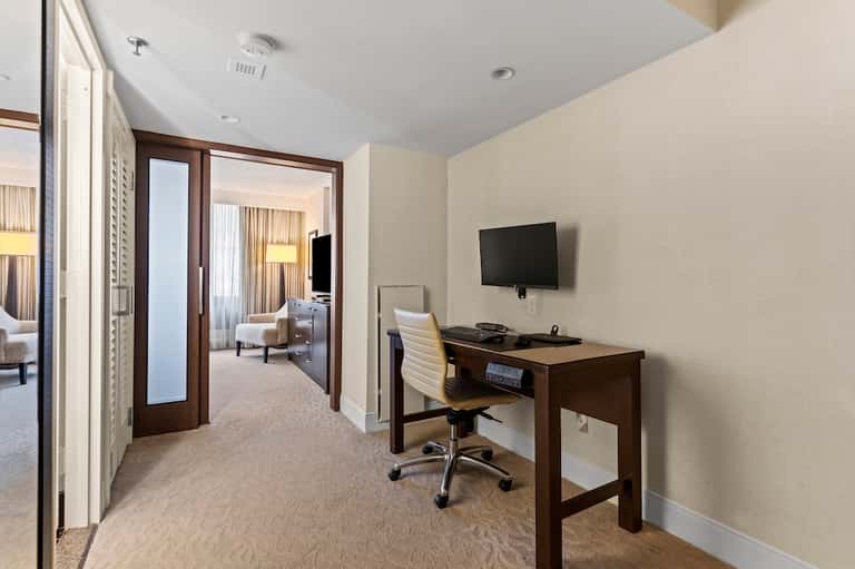 Hotel suite foyer with dark wood doors, desk workspace, and view into bedroom area with neutral color scheme.