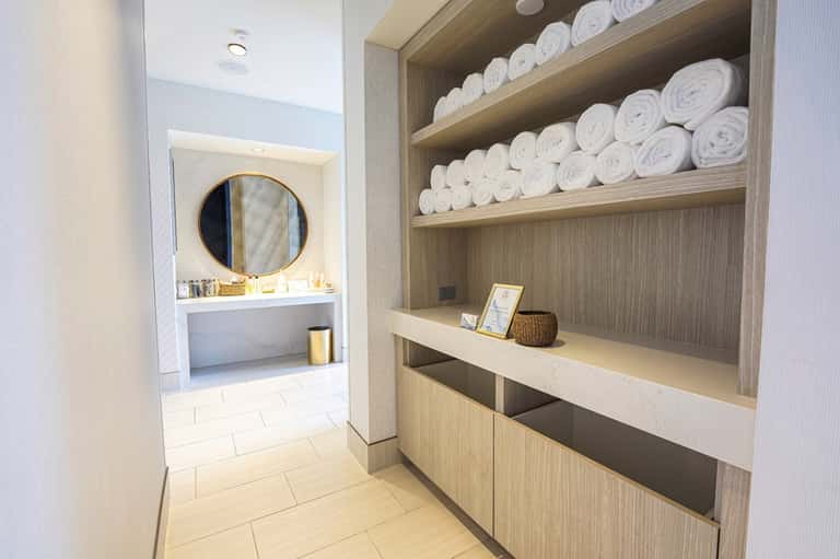 Spa hallway with built-in shelving displaying rolled white towels and vanity area with round mirror
