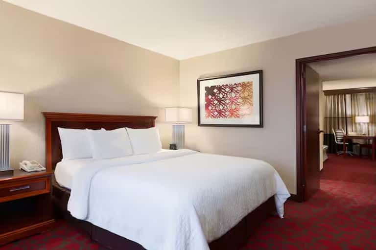 Hotel bedroom with queen bed, wood headboard, bedside lamps, and connecting suite