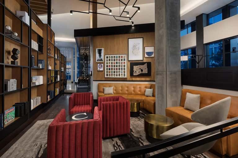 Modern hotel lounge with wooden shelving, red velvet chairs, orange seating, artwork, and floor-to-ceiling windows