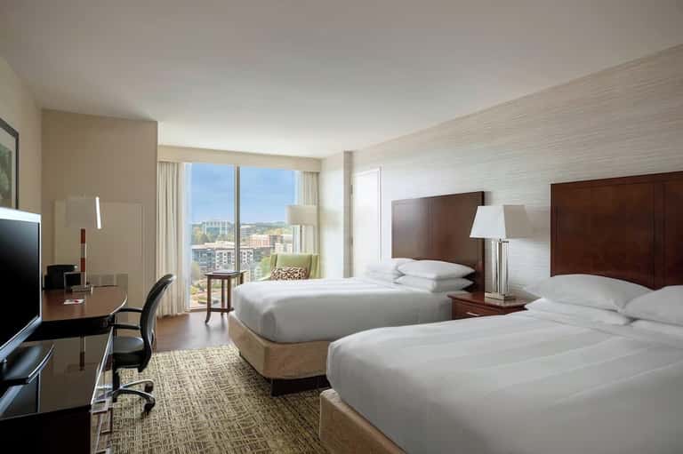 Modern hotel room with two queen beds, dark wood headboards, city view windows, and work desk with chair