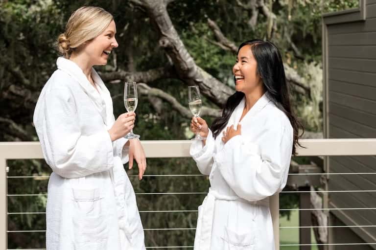 Two women in white spa robes toasting with champagne flutes in natural outdoor setting with trees