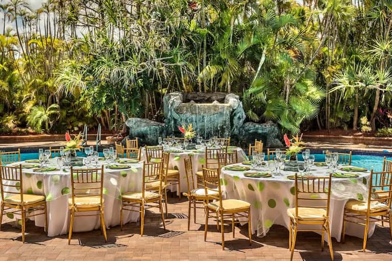 Elegant poolside banquet setup with white linens, gold chairs, and tropical waterfall backdrop