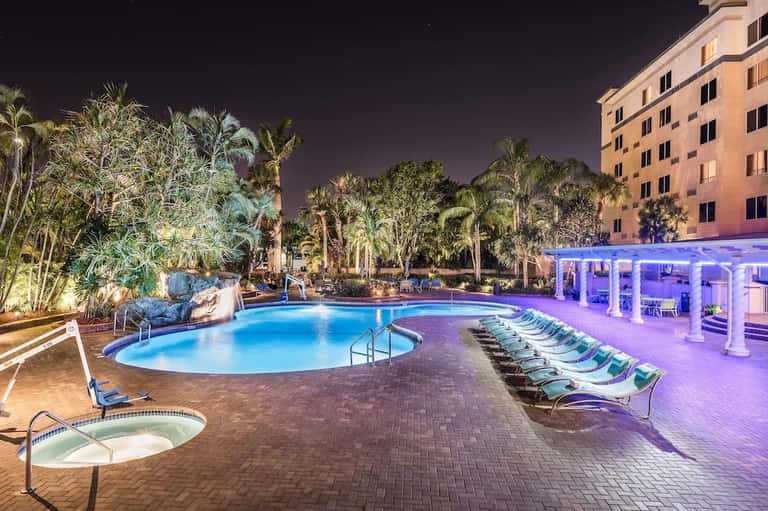 Illuminated resort pool at night with tropical landscaping, palm trees, and purple-lit cabana pavilion