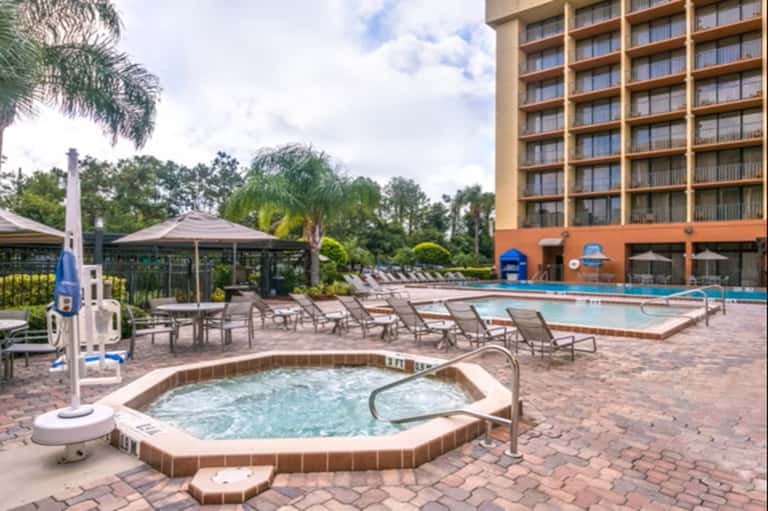 Resort pool and hot tub area with lounge chairs, gazebo, palm trees, and hotel building