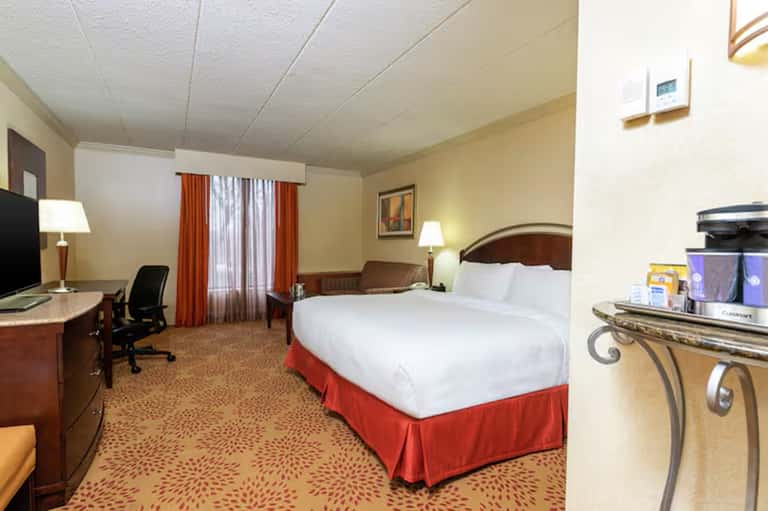 Hotel room with queen bed, red accents, desk with TV, and warm neutral color scheme