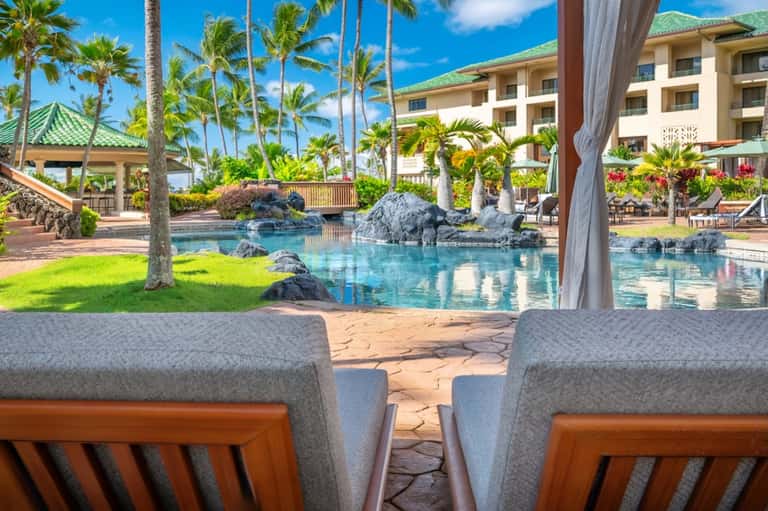 Tropical resort pool with palm trees, rock features, lounging chairs, and resort buildings