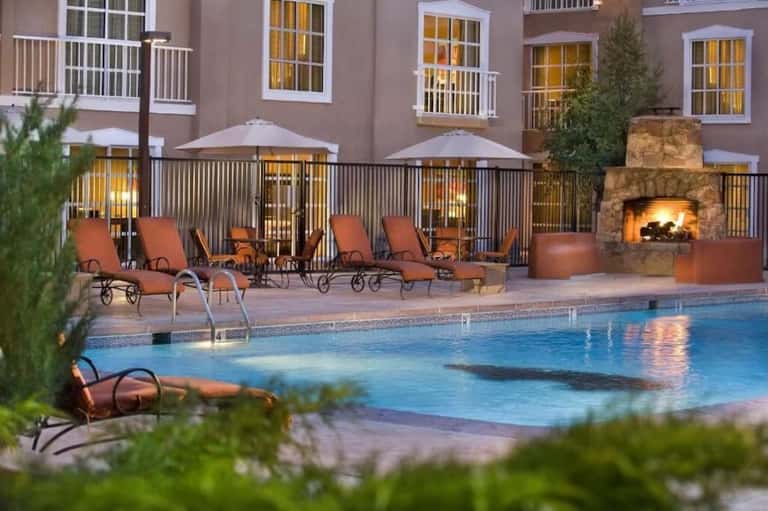 Resort pool at dusk with loungers, umbrellas, fireplace, building facade, and warm ambient lighting