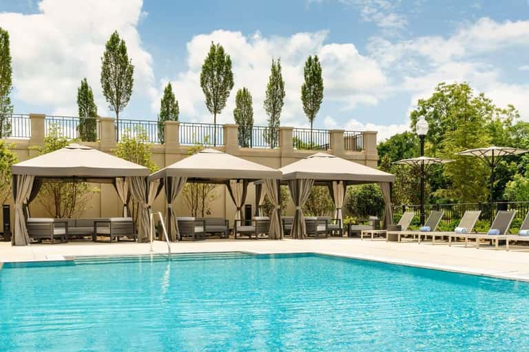 Resort pool with beige cabanas featuring curtains, tall cypress trees, and landscaped grounds