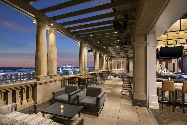 Upscale rooftop terrace at dusk with columns, lounge seating, bar area, and city skyline views