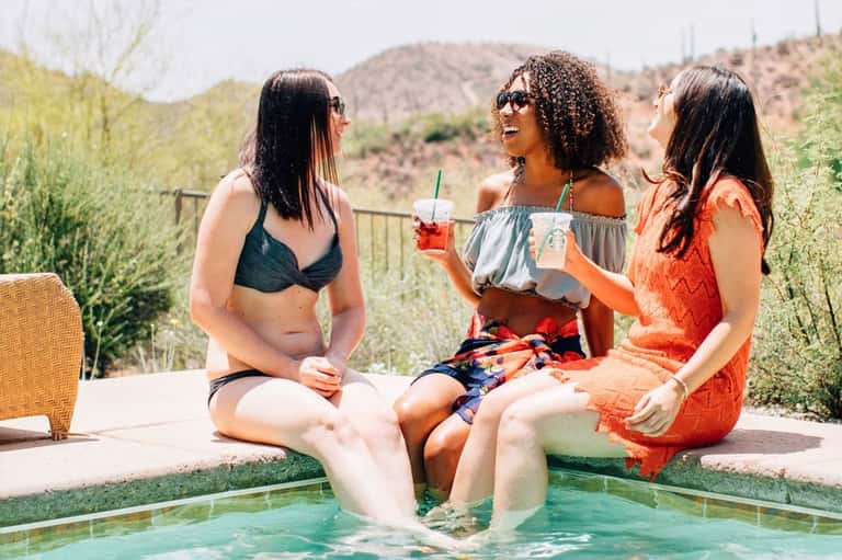 Three women sitting poolside with drinks, enjoying a desert landscape view