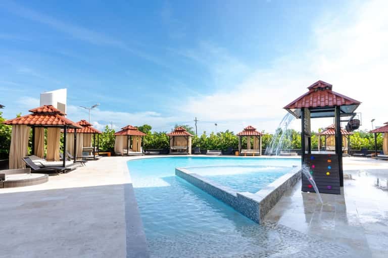 Resort pool area with multiple tiki-roofed cabanas, lounge chairs, and waterfall feature on sunny day