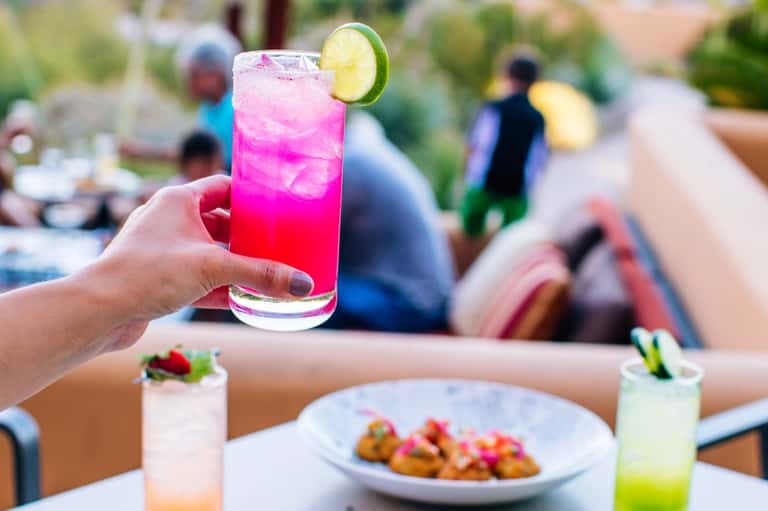 Hand holding vibrant pink cocktail with lime garnish at poolside gathering with food and guests