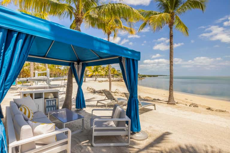 Beach cabana with blue curtains, lounge seating, mini fridge, palm trees, and ocean view