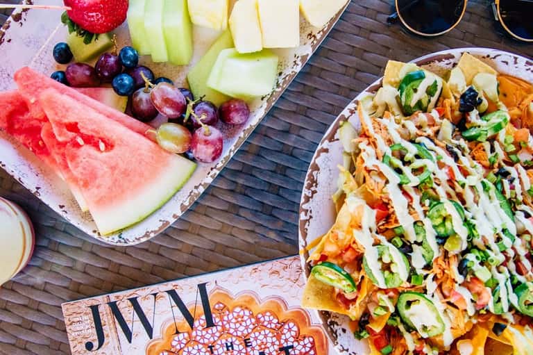 Overhead view of poolside spread with sliced watermelon, grapes, fresh fruit, and loaded nachos on wooden deck