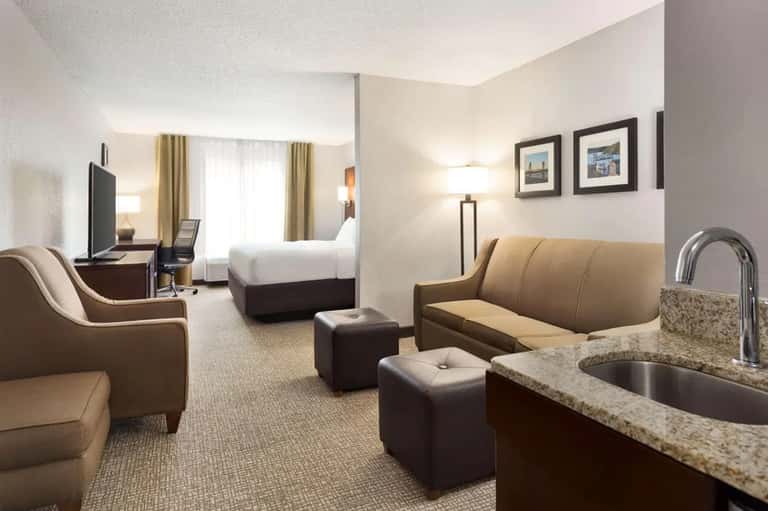 Spacious hotel suite featuring bed, sitting area with tan leather sofa, kitchenette with granite countertop, and flat-screen TV.