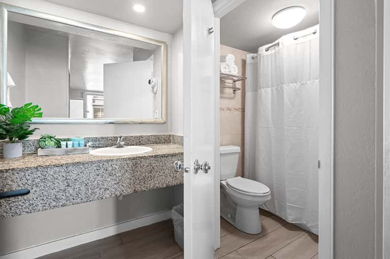 Clean hotel bathroom with granite countertop, large mirror, toilet, and shower with white curtain