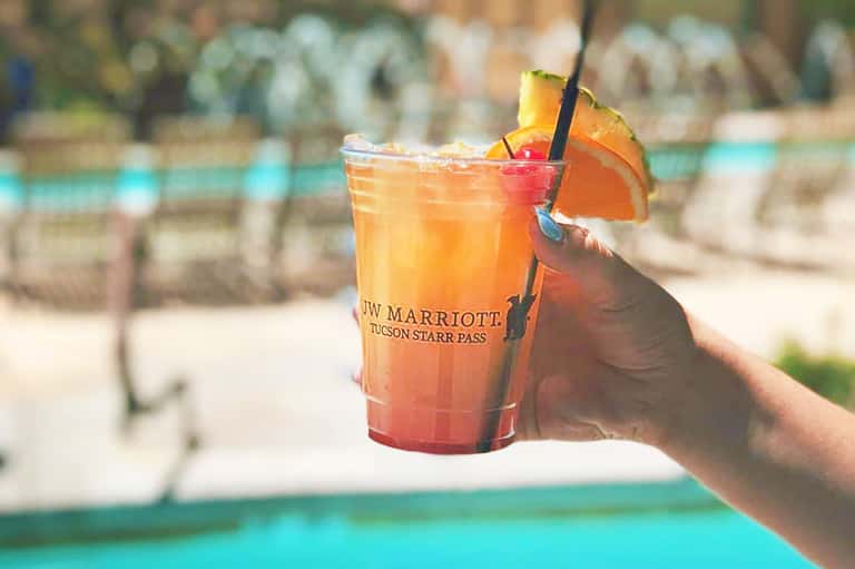 Hand holding a colorful cocktail with orange garnish by a resort pool