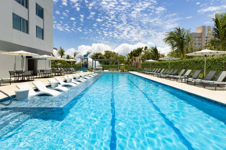 Resort lap pool with lane markers, white lounge chairs, umbrellas, and palm trees surrounding