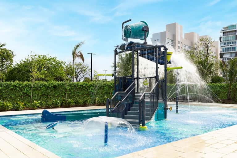 Water park splash pad with large bucket, blue water features, landscaping, and residential buildings