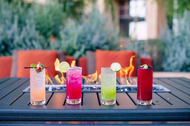 Five colorful cocktails lined up on a dark table with a fireplace and red seating in the background