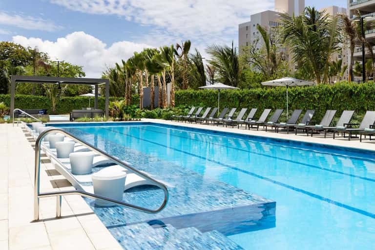 Resort lap pool with diving platform, white umbrellas, loungers, and palm tree landscaping