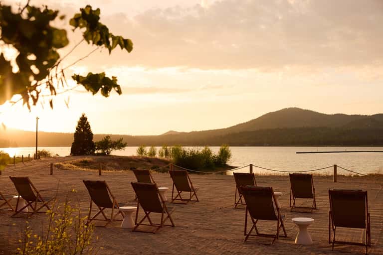 Beach chairs lined on sandy shore at sunset with lake, mountains, and pine trees