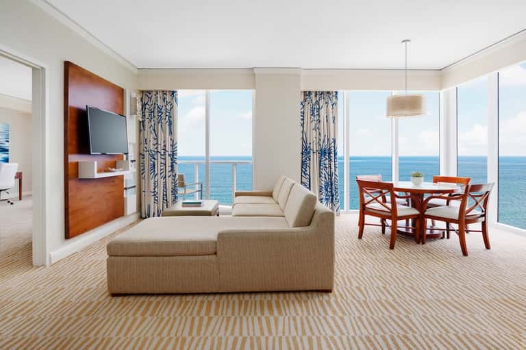 Upscale oceanfront suite with beige sectional sofa, ocean views, dining area, and patterned drapery