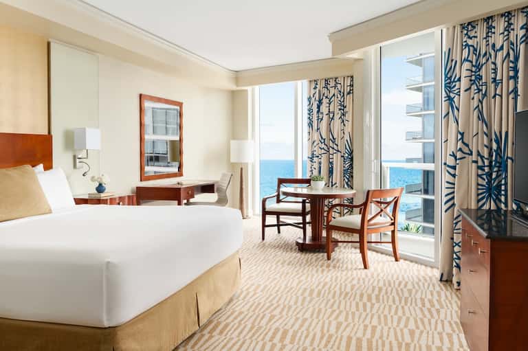 Luxury oceanfront hotel suite with king bed, sitting area, ocean view balcony, and patterned curtains