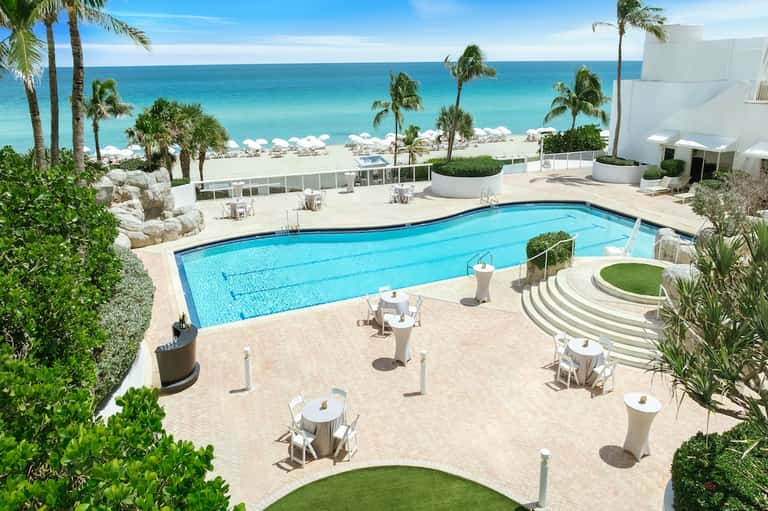 Beachfront resort pool overlooking turquoise ocean with white umbrellas and palm trees