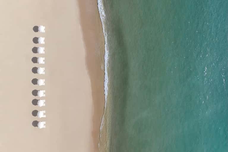 Aerial view of pristine sandy beach with rows of white umbrellas creating geometric shadows in turquoise shallow water