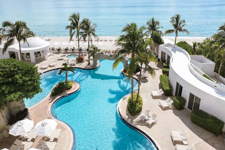 Beachfront resort with lagoon-style pool, white structures, palm trees, and ocean view