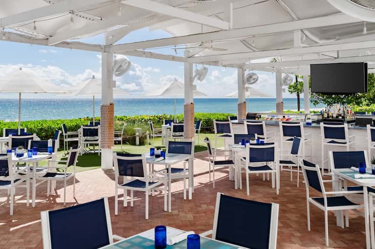 Beachfront restaurant terrace with white pergola, ocean view, dining tables, green landscaping
