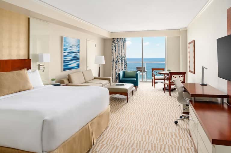 Luxury oceanfront hotel room with king bed, sofa seating, floor-to-ceiling windows, and sea views