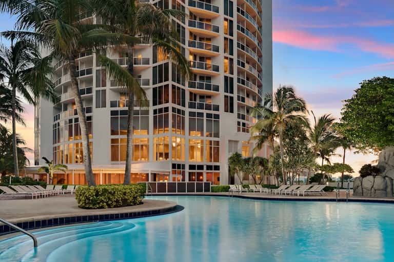 Modern coastal resort tower at dusk with palm trees and illuminated pool in foreground