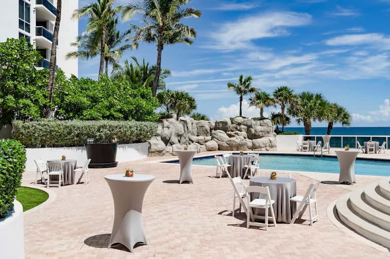 Poolside event space with white high-top tables, ocean view, and palm trees