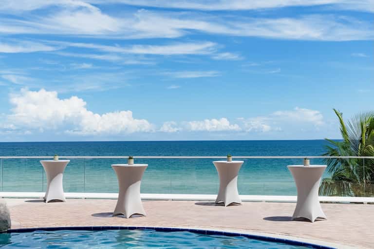Beachfront pool deck with white tables facing turquoise ocean and palm trees