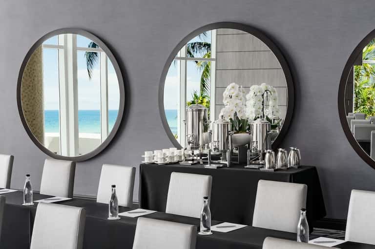Beachfront meeting room with large round mirrors, black conference table, white chairs, and ocean views