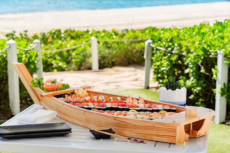 Beach setting with wooden boat-shaped food display, appetizers, ocean view, white fence, and green landscaping