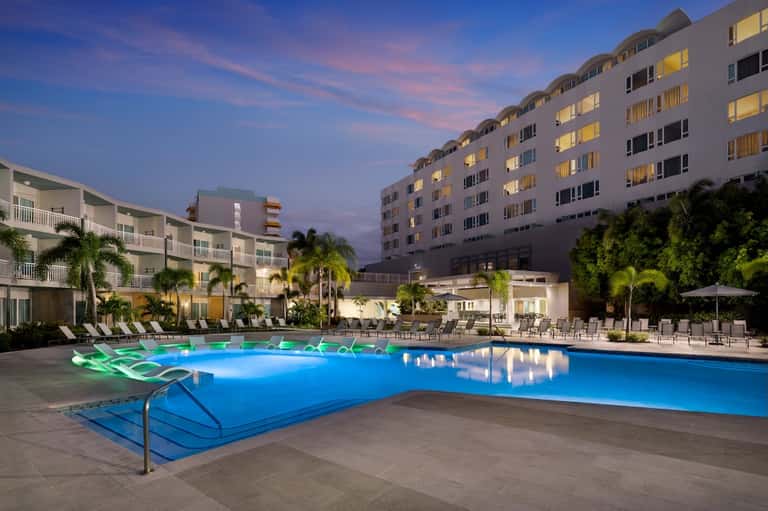 Illuminated resort pool at dusk with lounge chairs and multi-story hotel buildings