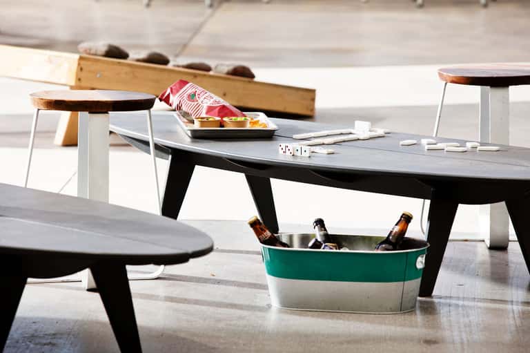 Modern lounge area with black tables, wooden stools, snacks, beverages in teal ice bucket, and playing cards on concrete floor