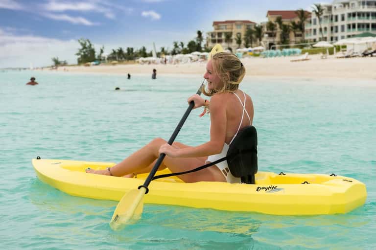Woman kayaking in clear turquoise ocean water with beachfront resort buildings visible in background