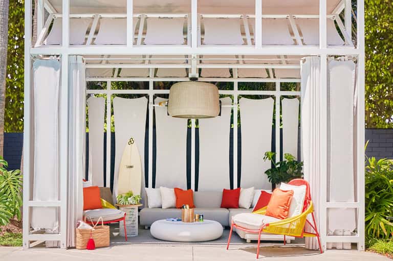 Modern pergola lounge with gray sofa, red cushions, hanging pendant light, and surfboard decor