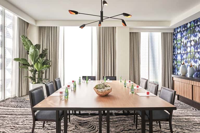 Modern conference room with long wooden table, gray upholstered chairs, floor-to-ceiling windows, and contemporary chandelier