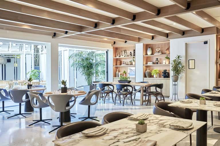 Modern restaurant dining area with marble tables, curved chairs, plants, and glass doors