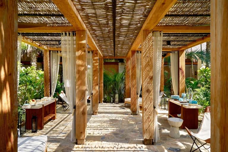 Covered spa pavilion with wooden pergolas, flowing curtains, red vessels, and tropical plants