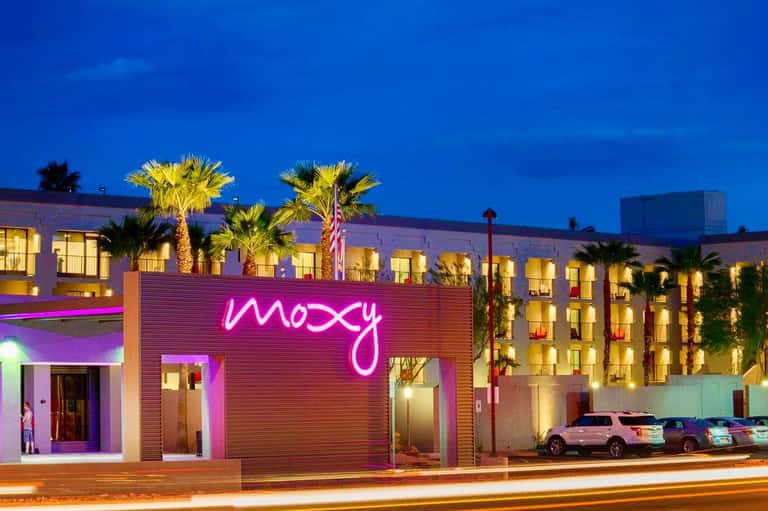 Modern hotel exterior at dusk with neon 'moxy' sign, illuminated palm trees, and parked cars