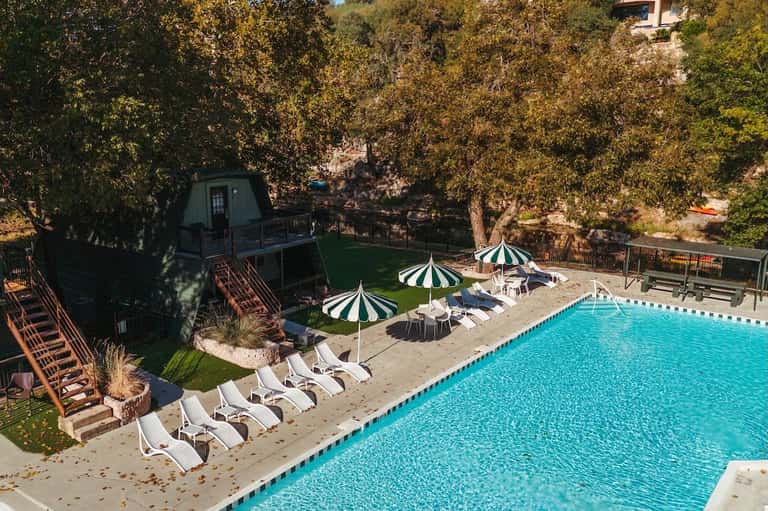 Resort pool surrounded by white loungers, striped umbrellas, and dense forest in natural setting